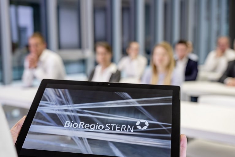 A person is holding a tablet, which shows the logo of the BioRegio STERN Management GmbH. The person is presenting in front of a group which is sitting in the background.