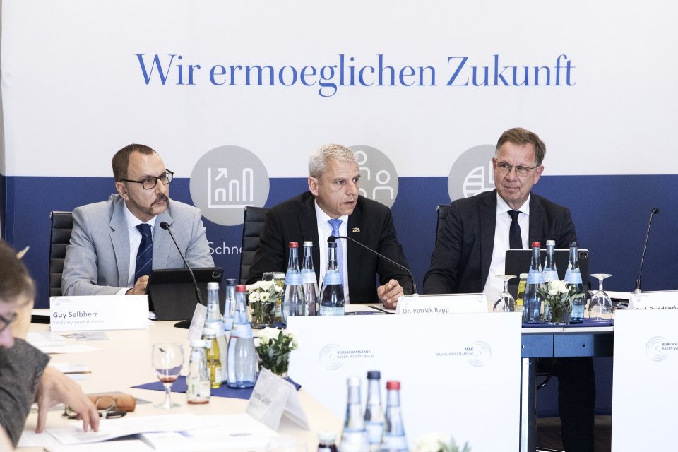 Drei Mönner sitzen nebeneinander an einem Tisch und halten eine Pressekonferenz.