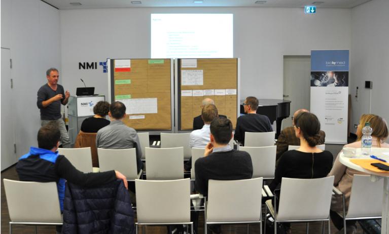 Mehrere Personen sitzen in einem kleinen Auditorium. Ein Teilnehmer steht neben zwei Flip-Charts und hält einen Vortrag.