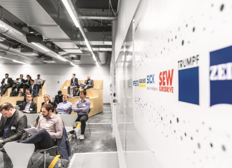 Mehrere Menschen sitzen in einem Podium. Auf der rechten Bildseite ist eine Wand mit Logos aufgestellt.