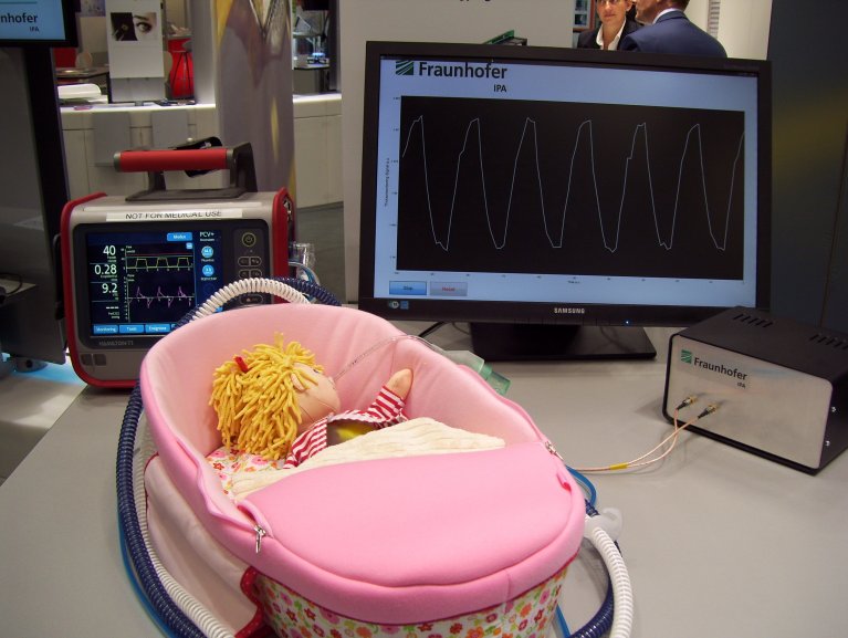 In einem kleinen rosa Bettchen liegt eine Puppe mit blonden Haaren, die einen Schlauch im Mund hat. Hinter der Puppe befinden sich zwei Monitore, welche verschiedene Kurven anzeigen.