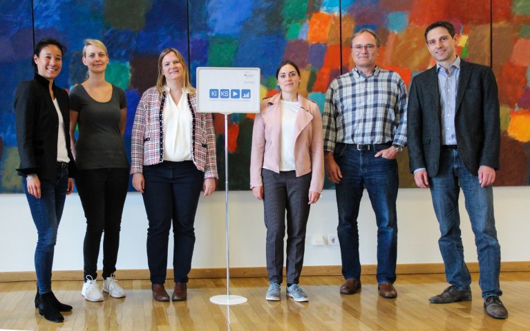 Mehrere Personen, die Projektpartner von KIKS, stehen in einer Gruppe. In der Mitte steht ein kleiner Aufsteller mit dem Logo des Förderprojekts KIKS.