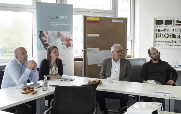 Four people sit at a table over a corner and listen to a lecturer.