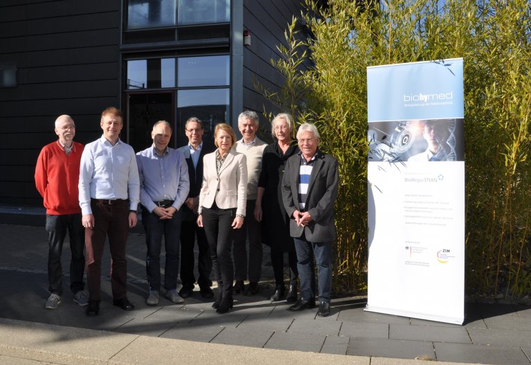 Die Partner es im Rahmen von biohymed geförderten Projekts PathChip stehen in einer Gruppe und sehen in die Kamera. Rechts von der Gruppe ist das Roll-up von biohymed aufgestellt.