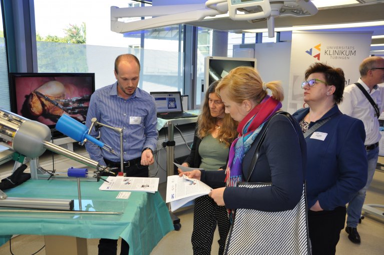 Four people are standing in front of a table with a green surgical drape and an apparatus and talk.