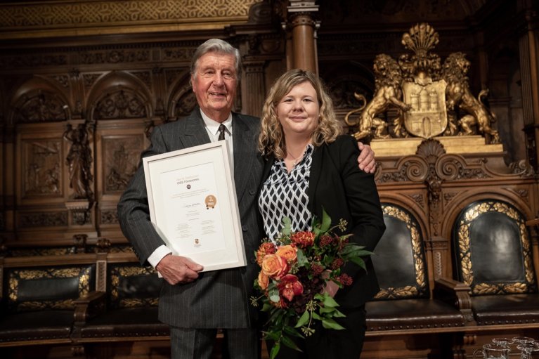 Eine Frau und ein Mann stehen nebeneinander. Der Mann hält eine Urkunde in den Händen und überreicht diese der Gewinnerin.