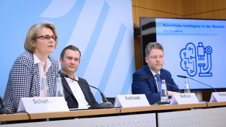 Drei Personen sitzen an einem länglichen Tisch zur Pressekonferenz. Hinter ihnen befindet sich ein großes Roll-up.