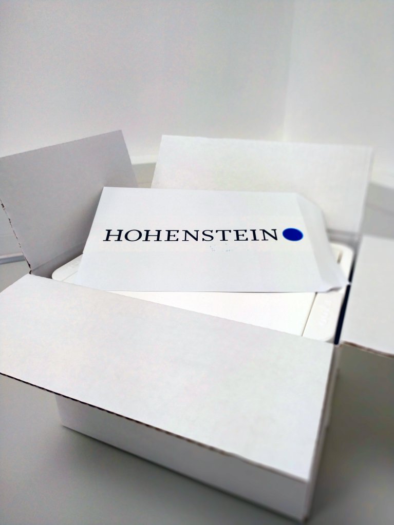 In einem aufgeklappten weißen Karton liegt ein weißer Umschlag mit der Aufschrift Hohenstein.