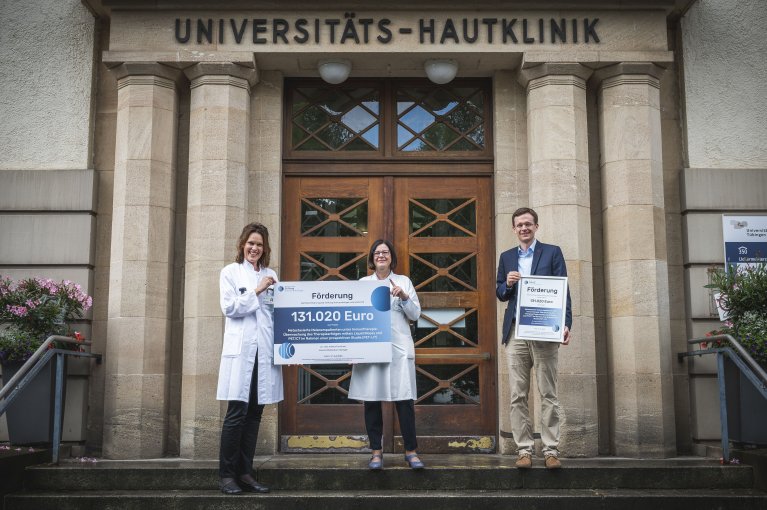 zwei Frauen im weißen Kittel halten einen großen Scheck in den Händen, ein Mann im Jackett präsentiert eine Urkunde