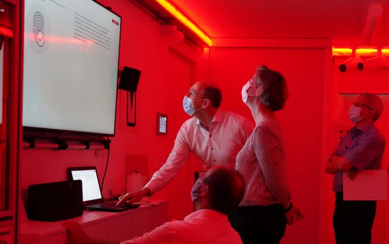 In einem rot beleuchteten Raum stehen mehrere Menschen und schauen gemeinsam auf eine Leinwand, auf welcher ein Fingerabdruck gezeigt wird.