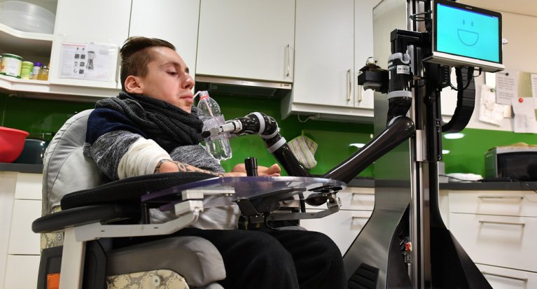Ein junger Mann sitzt in einem speziellen Stuhl und bekommt von einem Roboterarm eine Flasche gereicht aus der er trinken kann.