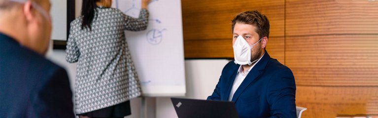 Ein Mann sitzt vor einer holzverkleideten Wand und trägt eine weiße Mund-Nasen-Bedeckung.