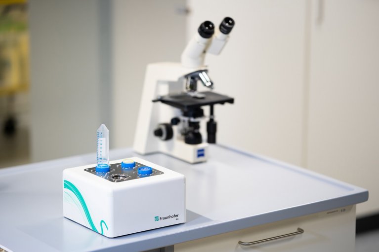 Ein viereckiges weißes Gerät steht auf einem Tisch. Das Gerät hat Platz für vier Proben. Im Hintergrund steht ein Mikroskop.