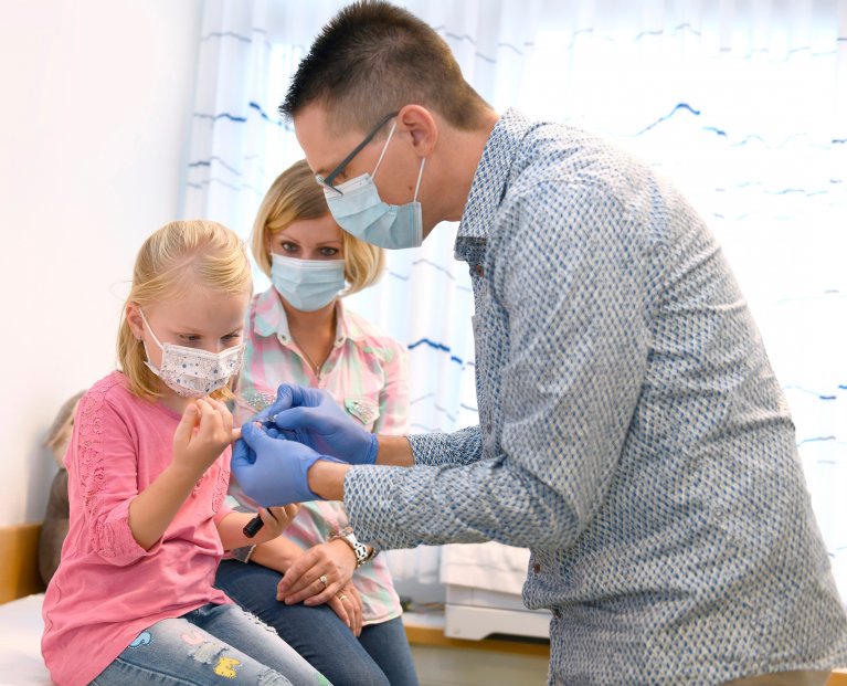 Ein Mann mit Mundschutz und Handschuhen misst bei einem kleinen blonden Mädchen den Blutzucker an einem Finger.