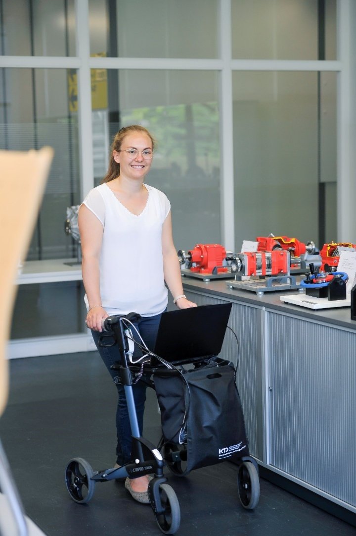 Eine junge Frau in weißem T-Shirt und Jeans schiebt einen Rollator in einem Gang.