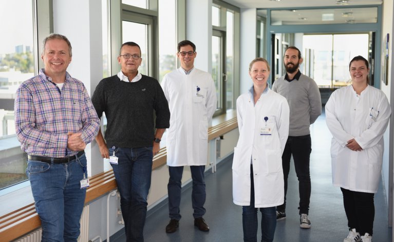 Sechs Personen stehen im hellen Gang eines Gebäudes und richten sich zur Kamera.