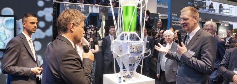 Dr. Robert Habeck ist links auf dem Bild zu sehen. Er hält ein Mikrofon in der Hand. Rechts auf dem Bild ist ein anderer Mann (Festo) zu sehen. Er hält ein Mikrofon und spricht. Zwischen beiden steht ein Modell (PhotoBionicCell von Festo)