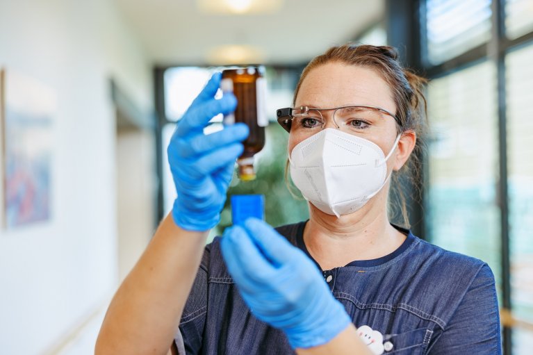 Frau mit Maske, Handschuhen füllt Flüssigkeit in eine kleine blaue Box. Sie trägt dabei eine AR-Brille.