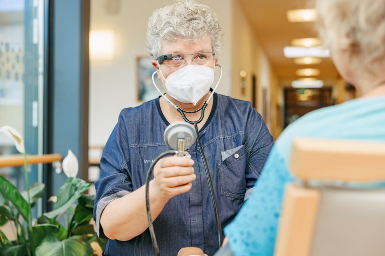 Ältere Frau hält ein Stethoskop in der Hand. Sie trägt eine AR-Brille. Vor ihr sitzt eine ältere Patientin, die von hinten gesehen wird.