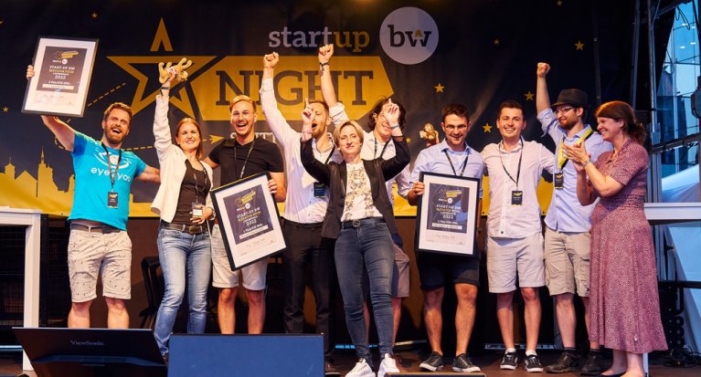 Gruppe von Menschen auf der Bühne. Im Hintergrund steht "startup bw NIGHT". Drei Personen halten Preise hoch. In der Mitte steht die Wirtschaftsministerin Dr. Nicole Hoffmeister-Kraut (BW)