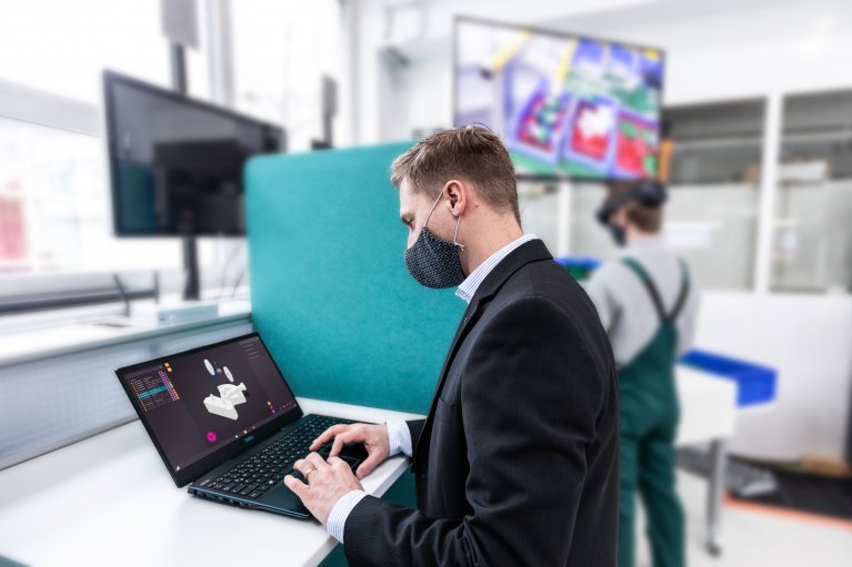 Im Vordergrund ist ein Mann in einem Anzug zu sehen. Er sitzt an einem Laptop. Auf dem Laptop scheint ein CAD-Programm zu laufen. Im Hintergrund steht ein Mann vor einem Fernseher, der an der Wand montiert ist.