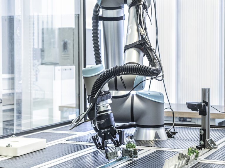 In der Mitte ist ein Robotiq-Roboter von weitem zu sehen. Mit seinem Roboterarm legt der Roboter Bauteile auf eine Metallschiene.