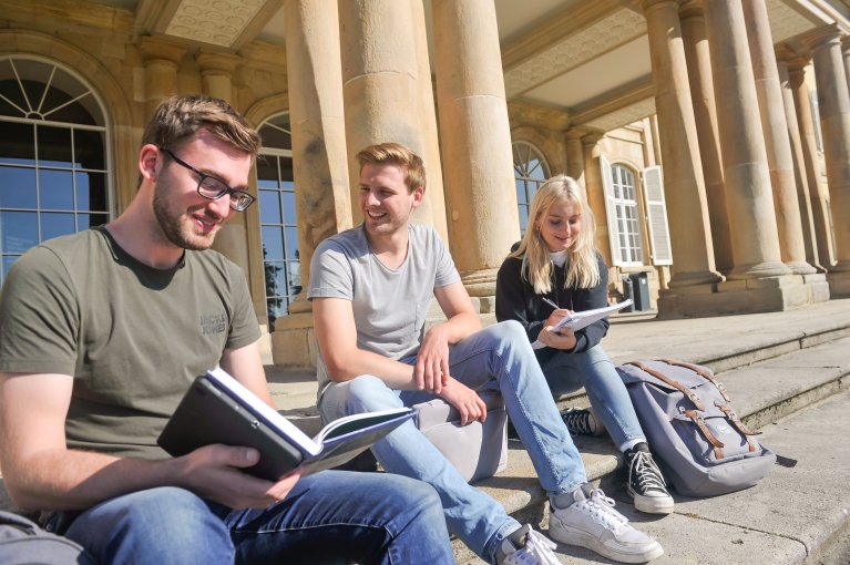 Three students are sitting on a staircase in front of a building with books in their hands.