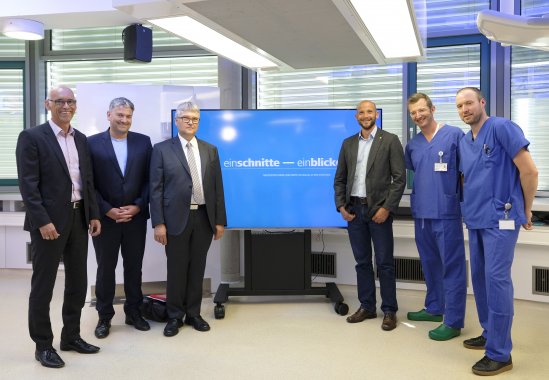 Sechs Männer stehen aufgereiht für ein Gruppenbild vor einem großen Bildschirm mit dem Logo von Einschnitte-Einblicke.