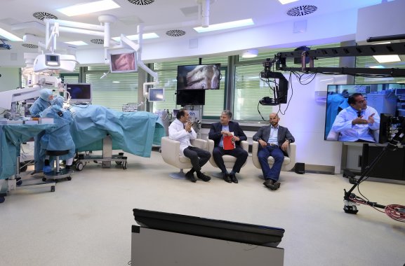 3 Männer sitzen in Sesseln im OP Saal der Klinischen Anatomie in Tübingen. Schräg hinter den Männern wird währenddessen operiert.
