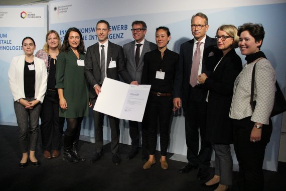 Acht Personen des KIKS Teams sowie Staatssekretär Dr. Ulrich Nussbaum stehen auf der Bühne und halten die Urkunde vor die Kamera.