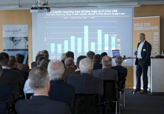 Dr. Siegfried Bialojan sthet vor einer Leinwand auf der die Präsentation abgebildet ist und richtet sich an das Publikum.