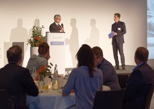 Klaus Eichenberg steht an einem Rednerpult und spricht in ein Mikrofon. Neben ihm steht der Moderator.