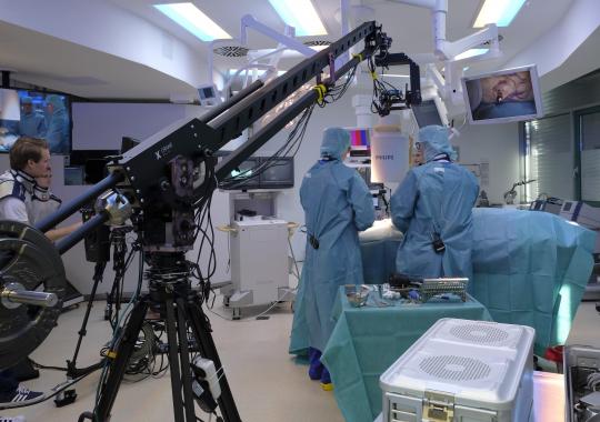 The people are in an operating room. A camera team transfers the operation with a large telescope camera to the auditorium.