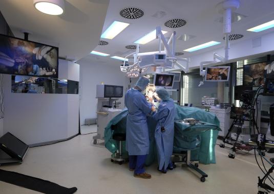 Two people are standing in an operating room and performing an operation on one hand. Several cameras are installed in the operating room, which transfer the operating room to the lecture hall.